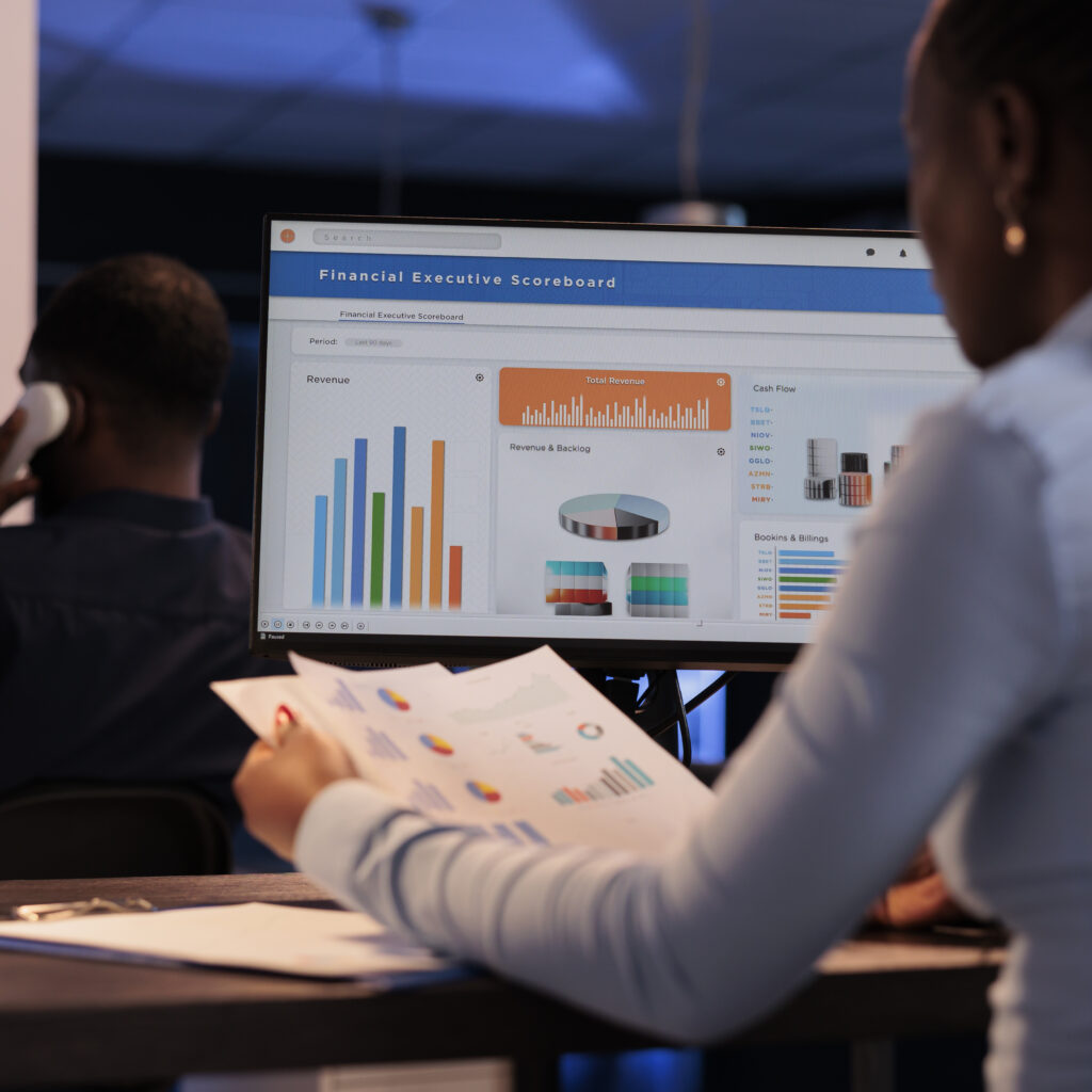 Young businesswoman working with statistics charts on computer monitor, analyzing company data on paperwork. Using online information to create research report before deadline, at night after hours.